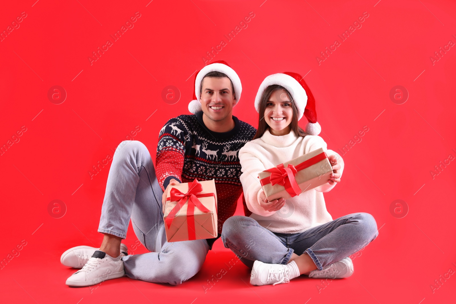 Photo of Beautiful happy couple in Santa hats and sweaters sitting with Christmas gifts on red background