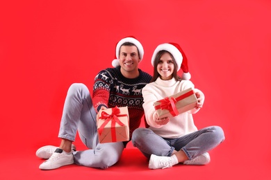 Beautiful happy couple in Santa hats and sweaters sitting with Christmas gifts on red background