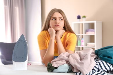 Emotional woman near board with iron and pile of clothes at home