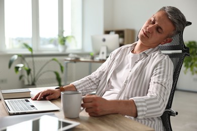 Man with cup of drink snoozing at wooden table in office