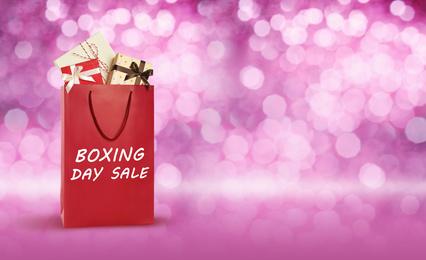 Image of Red shopping bag with text Boxing Day Sale full of gifts on blurred pink background, closeup. Space for text