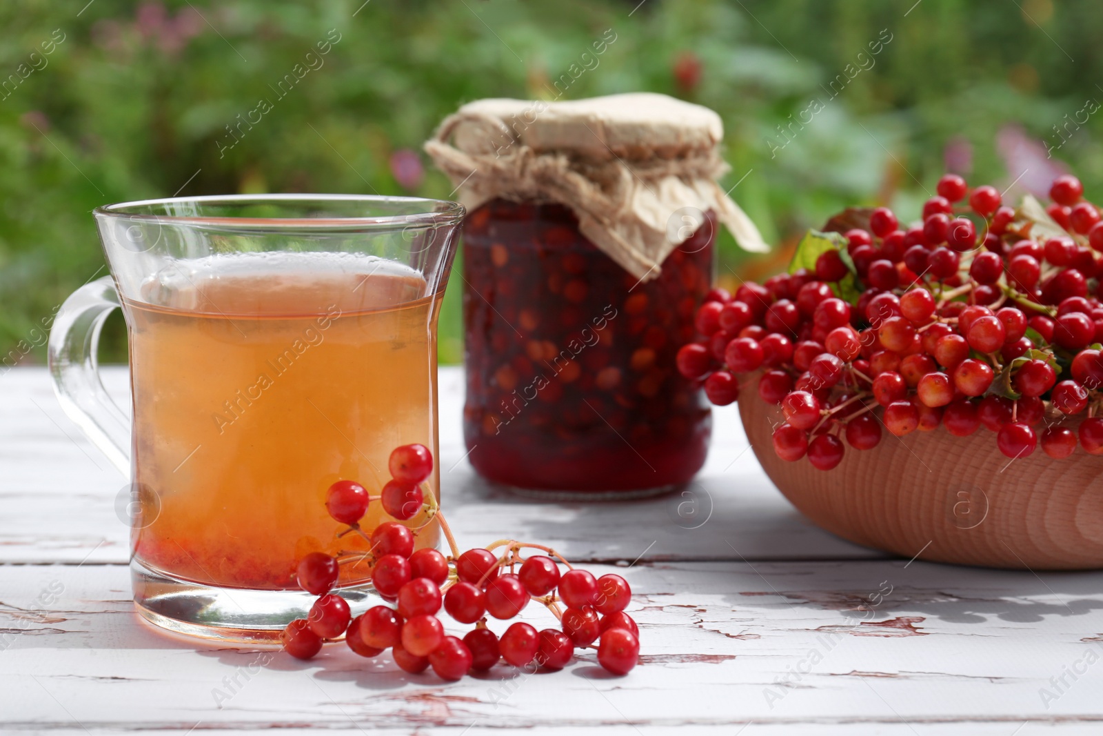 Photo of Cup of tea, jam and ripe viburnum berries on white wooden table outdoors