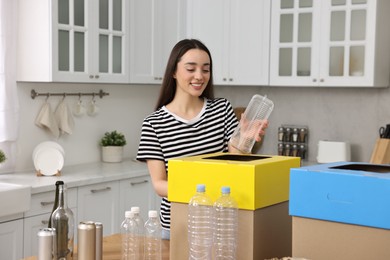 Photo of Garbage sorting. Smiling woman throwing plastic container into cardboard box in kitchen