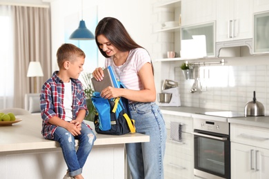 Happy mother putting textbooks into little child's school bag in kitchen