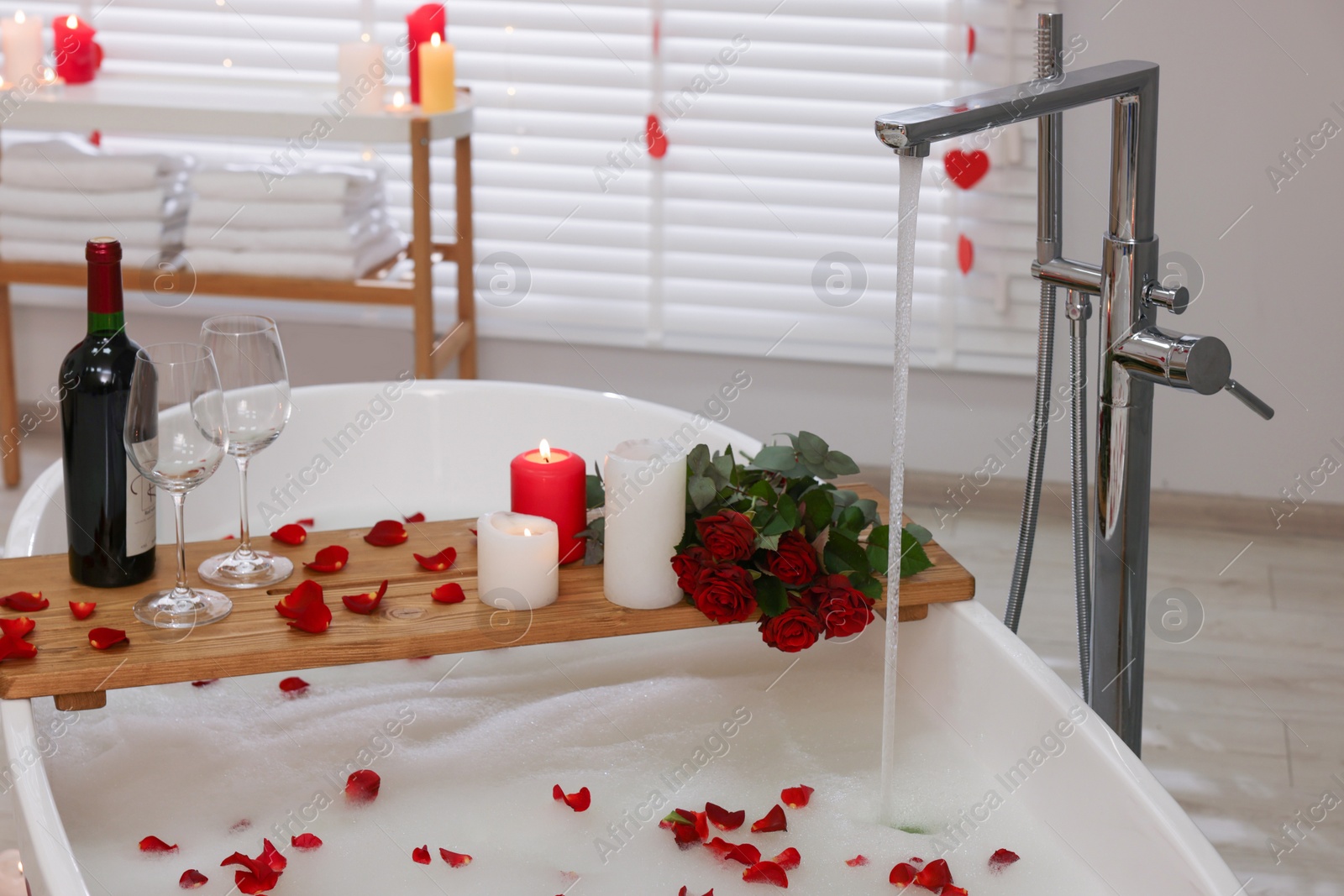 Photo of Wooden tray with wine, burning candles and rose petals on tub in bathroom. Valentine's day celebration