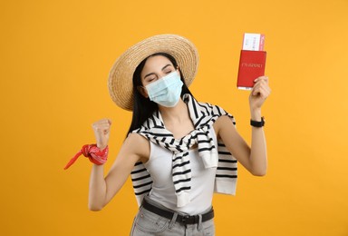 Female tourist in medical mask with ticket and passport on yellow background. Travelling during coronavirus pandemic