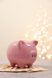 Photo of Piggy bank with euro banknotes on grey table against blurred lights, space for text