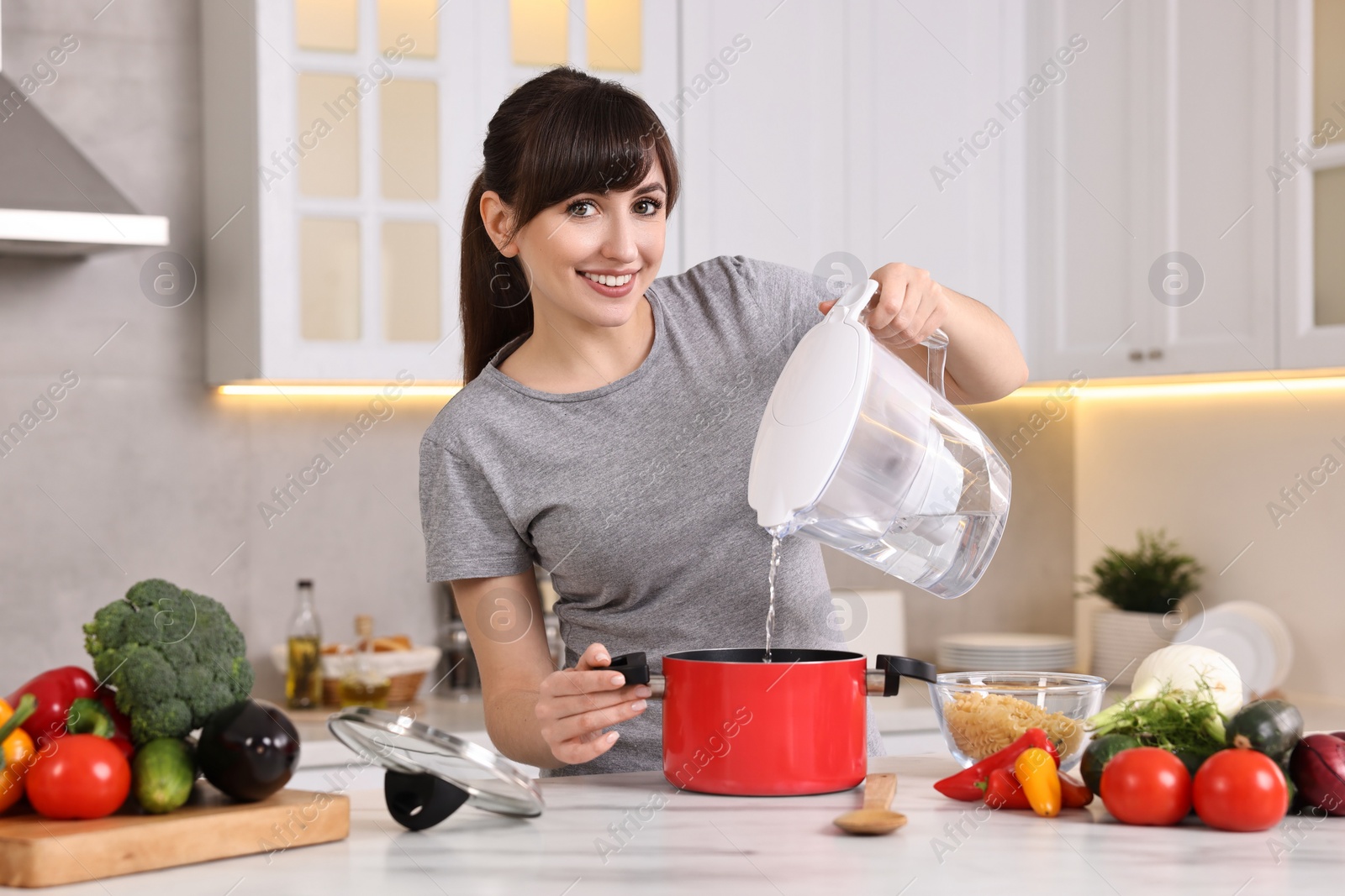 Photo of Happy young housewife pouring water in pot at white marble table in kitchen. Cooking process