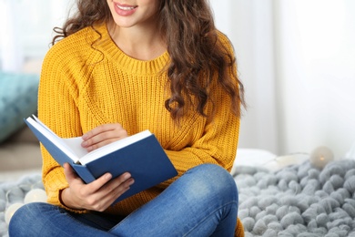 Photo of Young beautiful woman in warm sweater reading book on bed at home