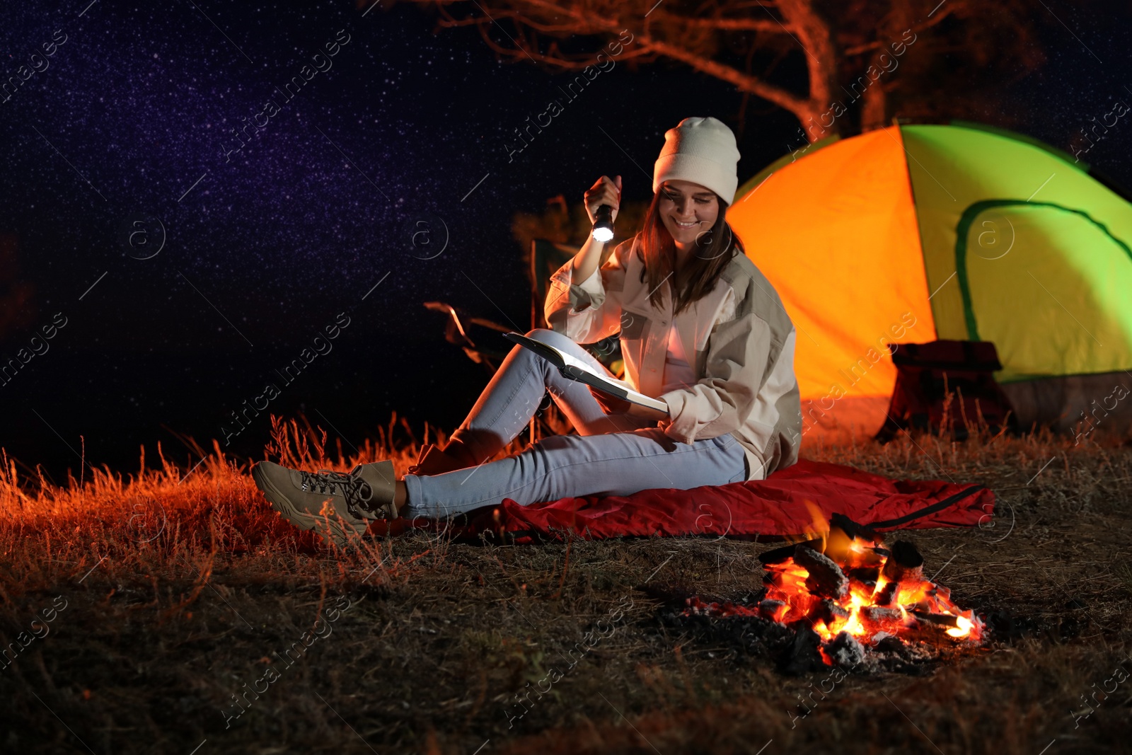 Photo of Young woman with flashlight reading book near bonfire at night. Camping season