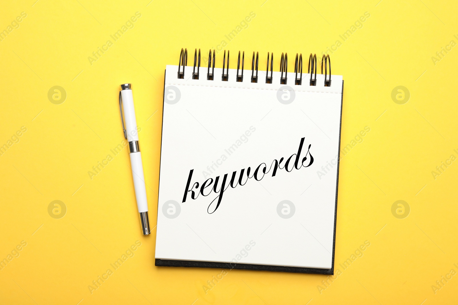 Image of Stylish notebook with text KEYWORDS and pen on yellow background, top view