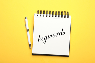 Image of Stylish notebook with text KEYWORDS and pen on yellow background, top view