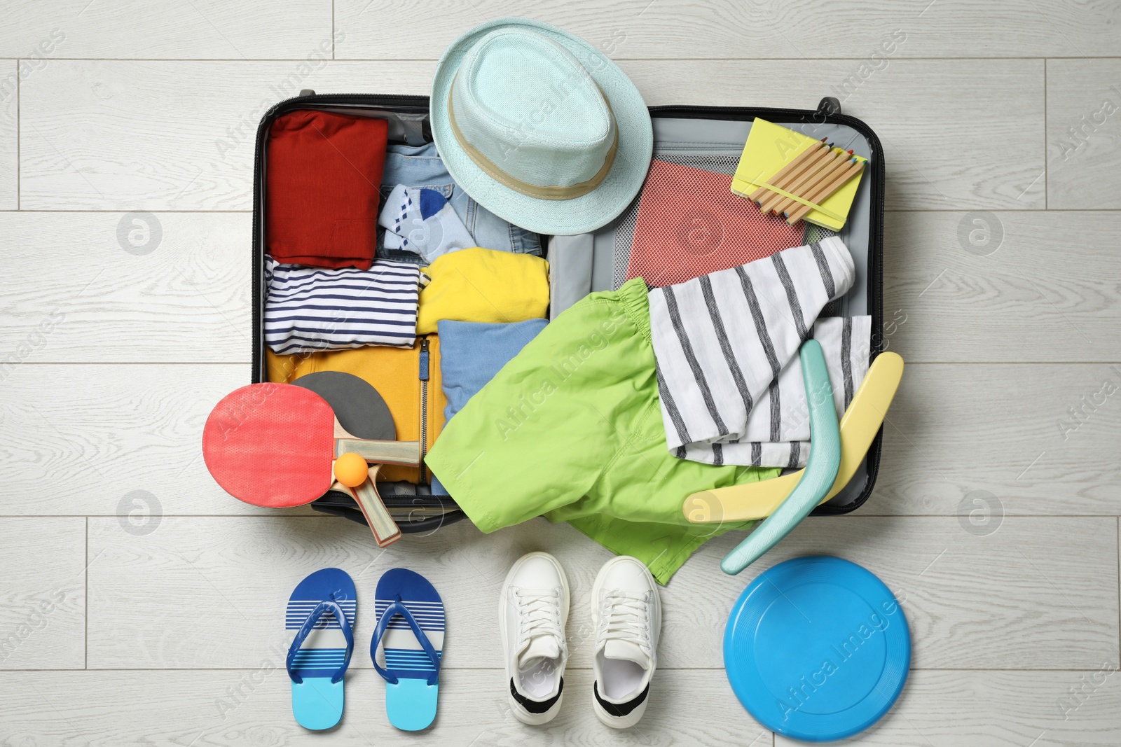 Photo of Open suitcase with stylish clothes, accessories, sport equipment and shoes on floor, flat lay. Summer vacation