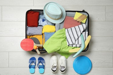 Open suitcase with stylish clothes, accessories, sport equipment and shoes on floor, flat lay. Summer vacation