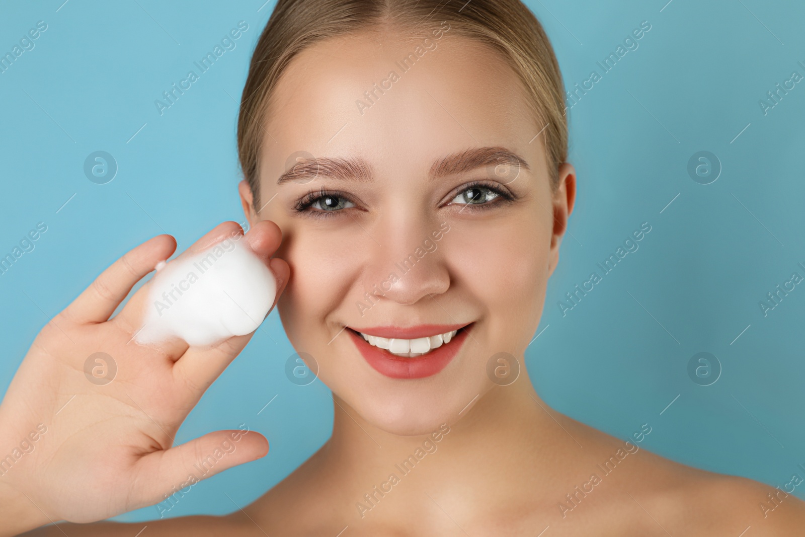 Photo of Young woman washing face with cleansing foam on light blue background. Cosmetic product