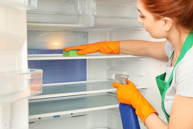 Woman in protective gloves cleaning refrigerator with sponge indoors