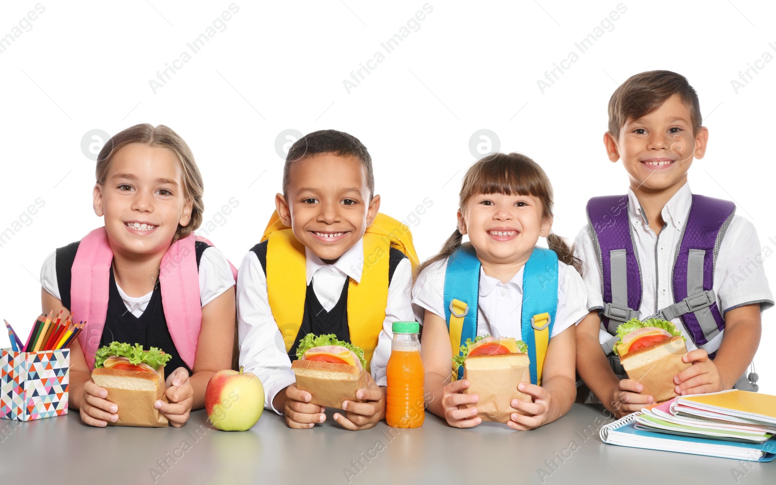 Photo of Schoolchildren with healthy food and backpacks sitting at table on white background