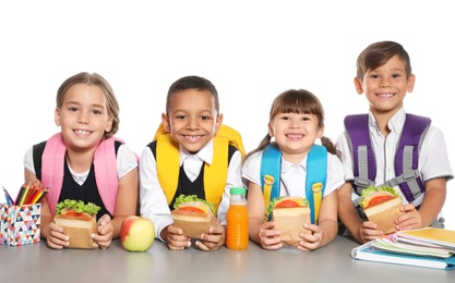 Schoolchildren with healthy food and backpacks sitting at table on white background