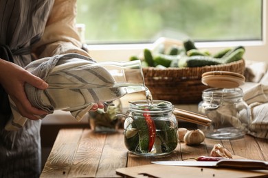 Photo of Woman pouring water into jar with cucumbers at wooden table, closeup. Pickling vegetables