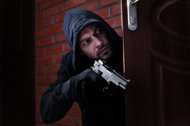 Man with gun spying behind open door indoors. Criminal offence