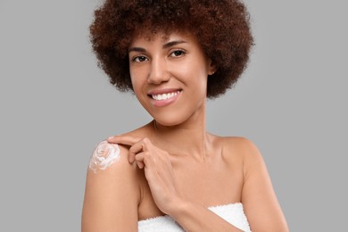 Beautiful young woman applying body cream onto shoulder on grey background