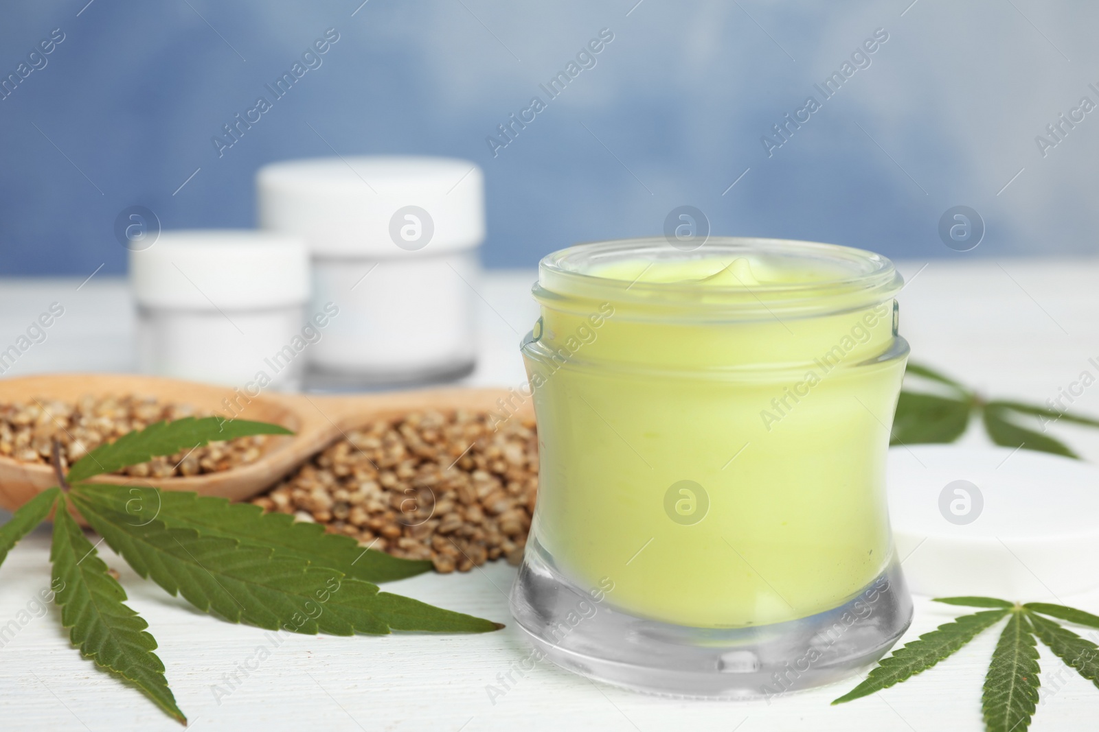 Photo of Jar of hemp cream and seeds on white wooden table against blue background, closeup. Organic cosmetics