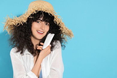 Beautiful young woman in straw hat holding tube of sun protection cream on light blue background, space for text