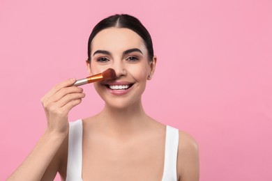 Beautiful woman applying makeup with brush on pink background. Space for text