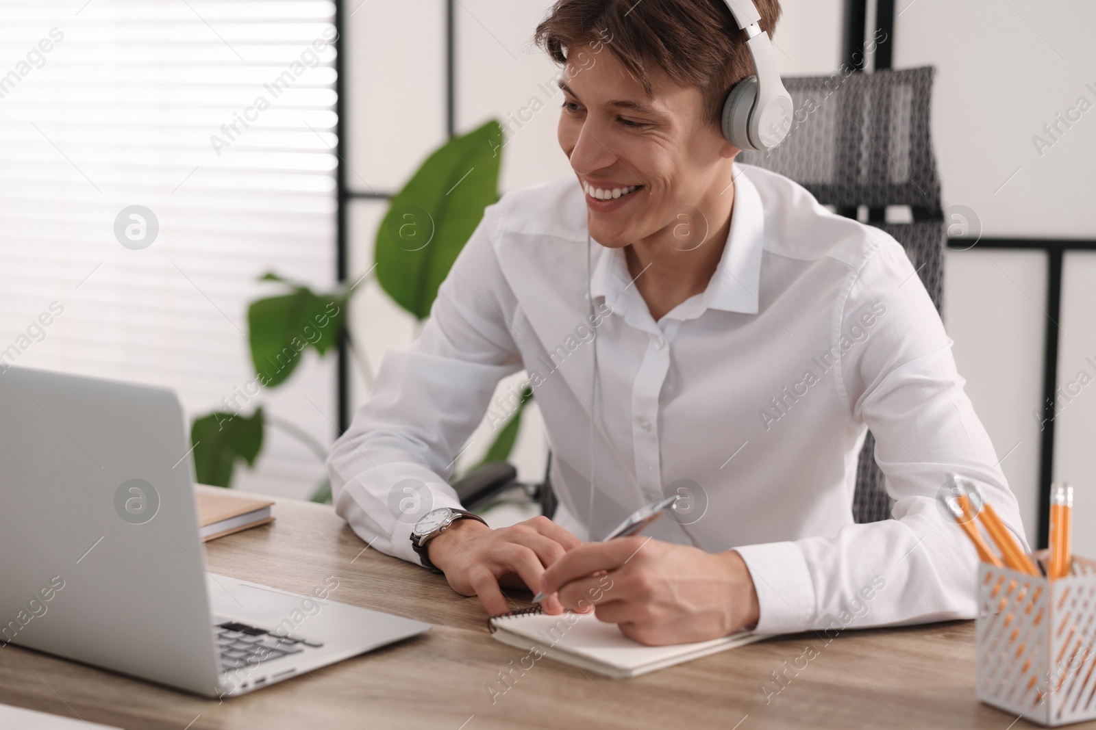 Photo of Man in headphones taking notes during webinar at wooden table indoors