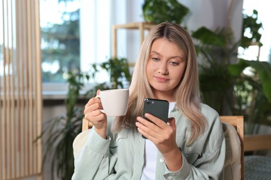 Photo of Woman with smartphone and cup of drink sitting in armchair at home