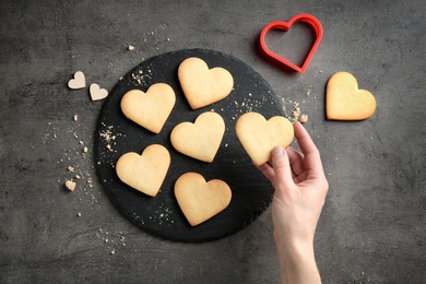 Woman holding homemade heart shaped cookie over table with pastry, top view