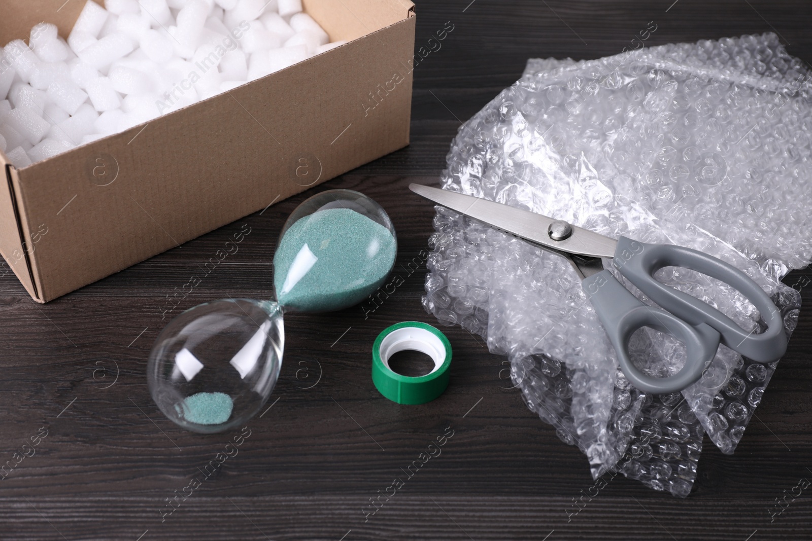 Photo of Sand hourglass, bubble wrap and packaging items on dark wooden table