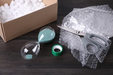 Photo of Sand hourglass, bubble wrap and packaging items on dark wooden table