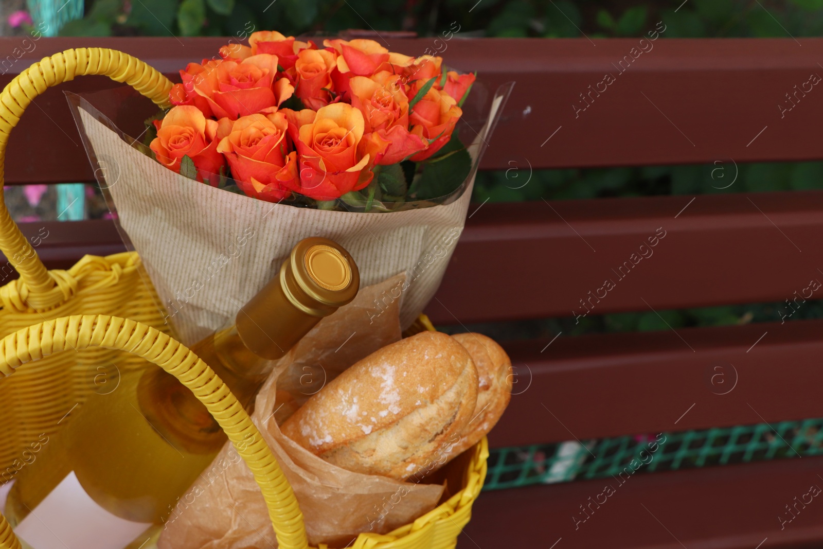 Photo of Beautiful roses, bottle of wine and baguettes in yellow wicker bag outdoors, closeup. Space for text