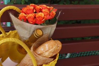 Photo of Beautiful roses, bottle of wine and baguettes in yellow wicker bag outdoors, closeup. Space for text