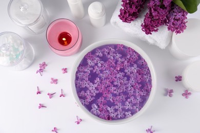Bowl of water with lilac flowers, spa products and burning candle on white table, flat lay