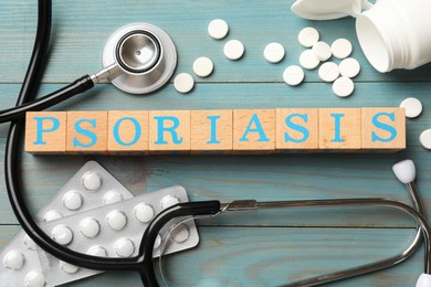 Word Psoriasis made of cubes with letters, stethoscope and pills on light blue wooden table, flat lay