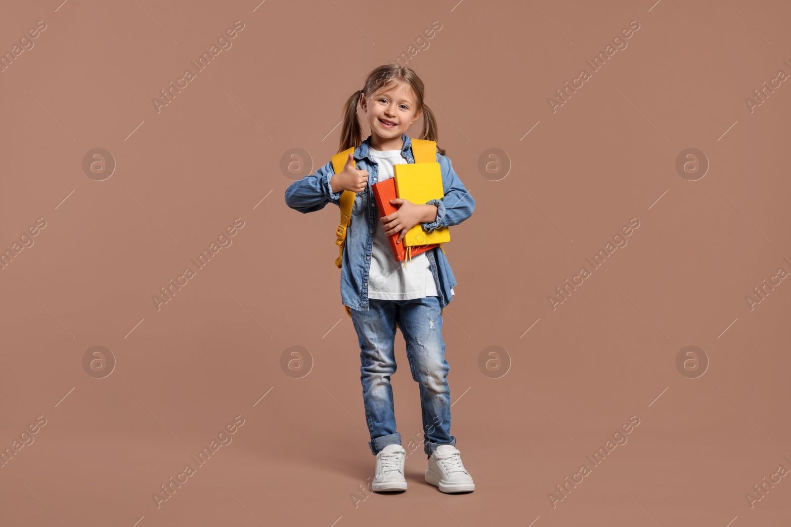 Photo of Happy schoolgirl with backpack and books showing thumb up gesture on brown background