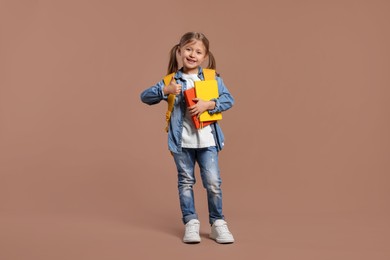 Photo of Happy schoolgirl with backpack and books showing thumb up gesture on brown background