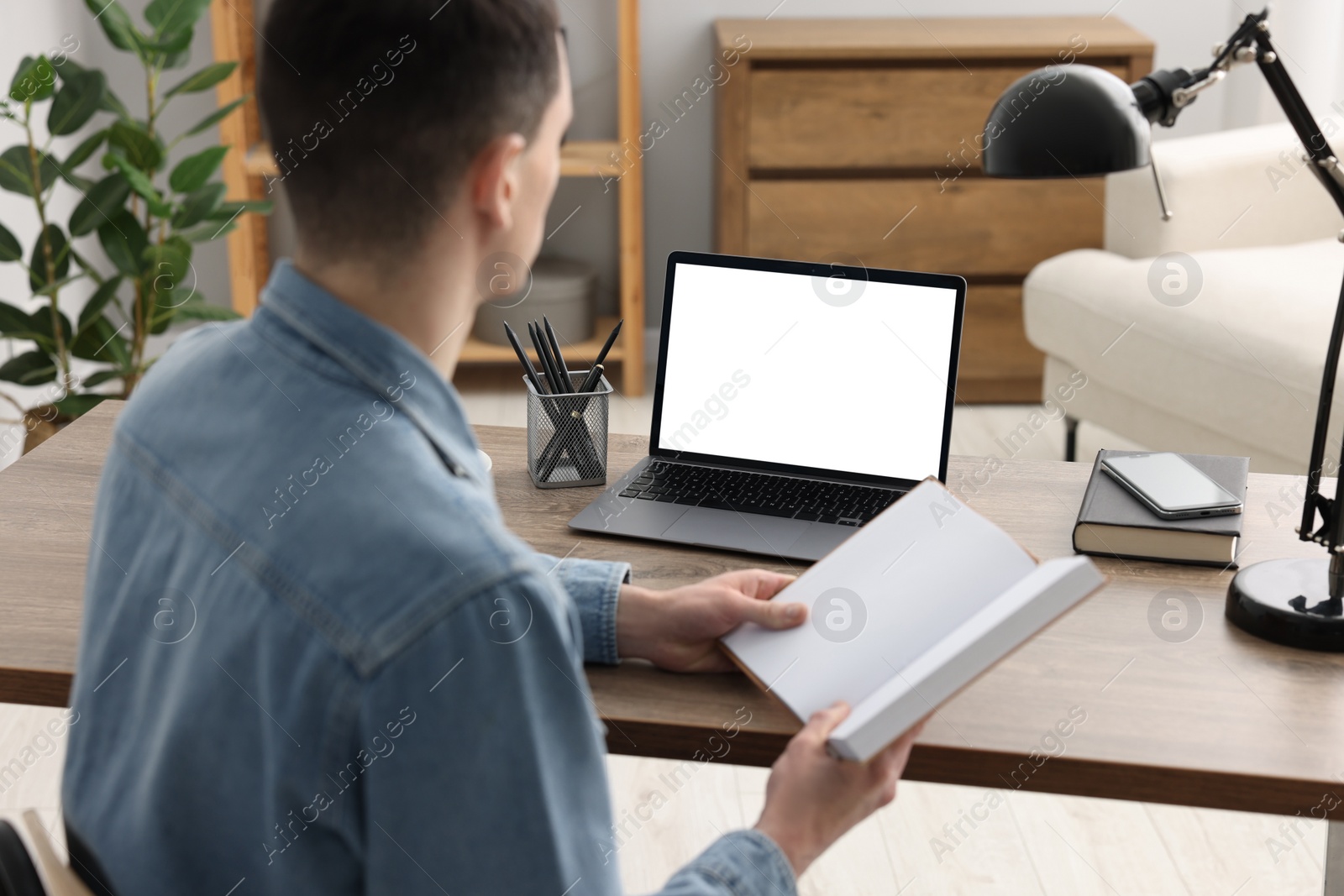 Photo of E-learning. Young man with book during online lesson at table indoors, closeup