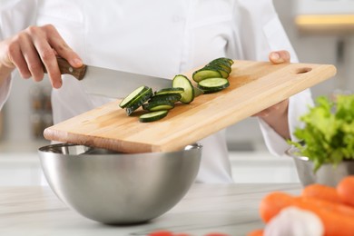 Photo of Professional chef putting cut cucumber into metal bowl at white marble table in kitchen, closeup