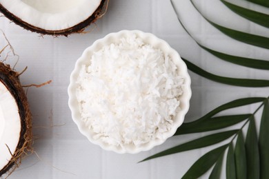 Coconut flakes in bowl, nuts and palm leaf on white tiled table, flat lay