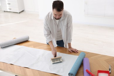 Man applying glue onto wallpaper sheet at wooden table indoors