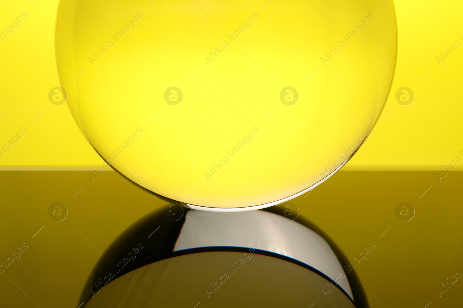 Photo of Transparent glass ball on mirror surface against yellow background, closeup