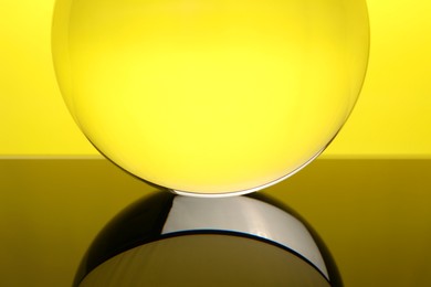 Transparent glass ball on mirror surface against yellow background, closeup