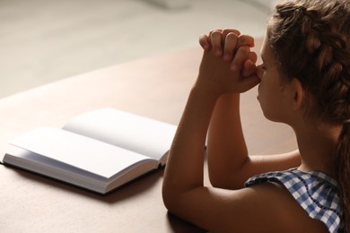 Photo of Cute little girl praying over Bible at table indoors, closeup