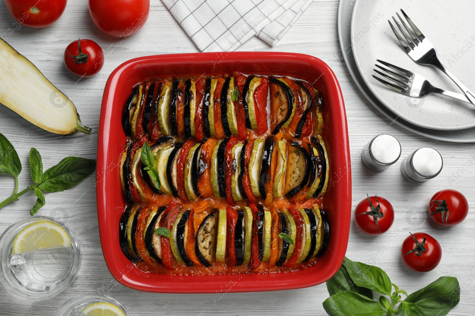Photo of Delicious ratatouille in baking dish and ingredients on white wooden table, flat lay