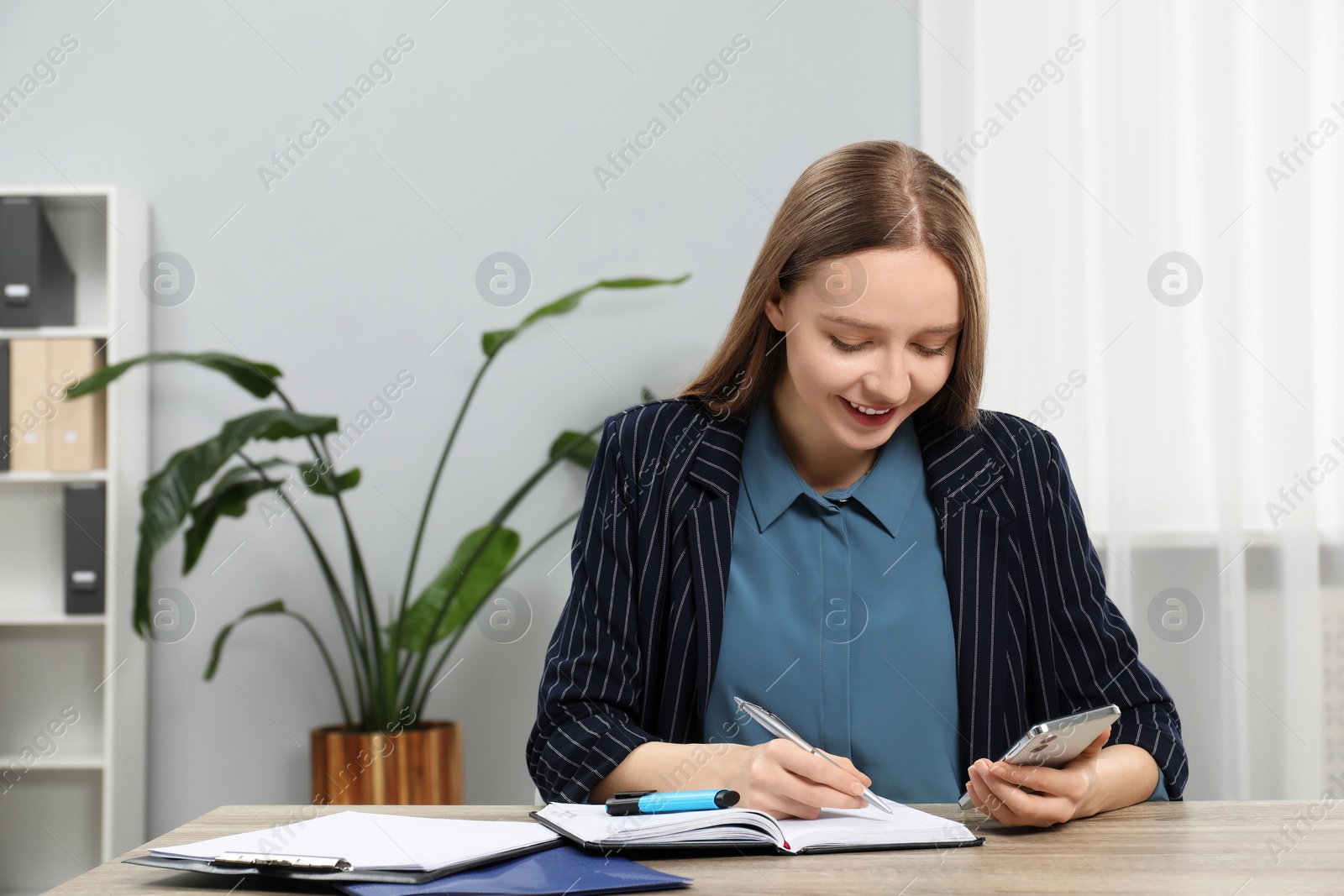 Photo of Woman taking notes while using smartphone at wooden table in office