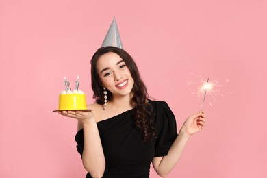 Photo of Coming of age party - 21st birthday. Smiling woman holding delicious cake with number shaped candles and sparkler on pink background