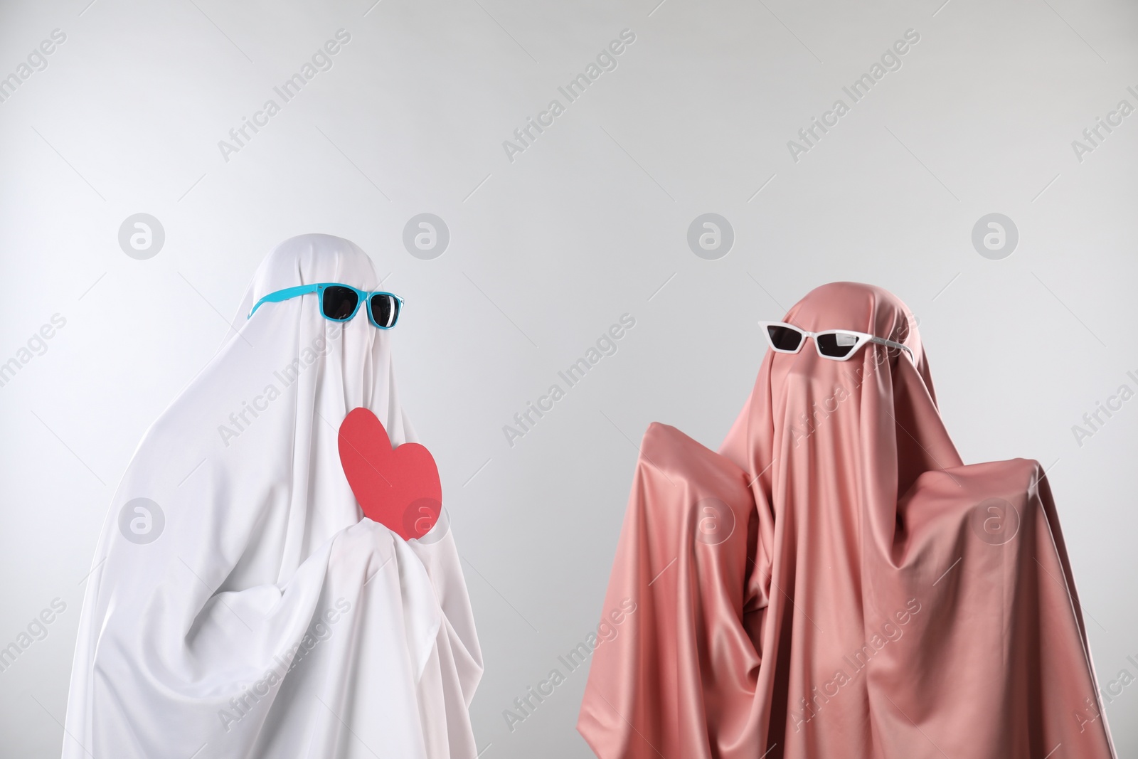 Photo of Cute ghosts. Couple in color sheets and sunglasses with red heart on light grey background